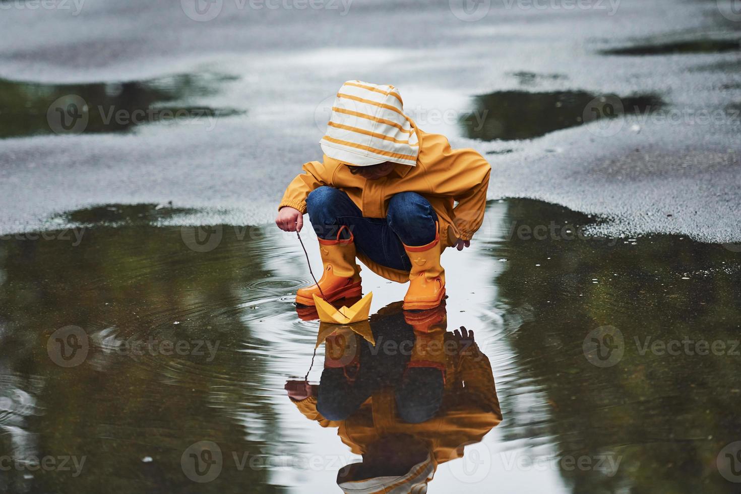 Kid in yellow waterproof cloak and boots playing with paper handmade boat toy outdoors after the rain photo