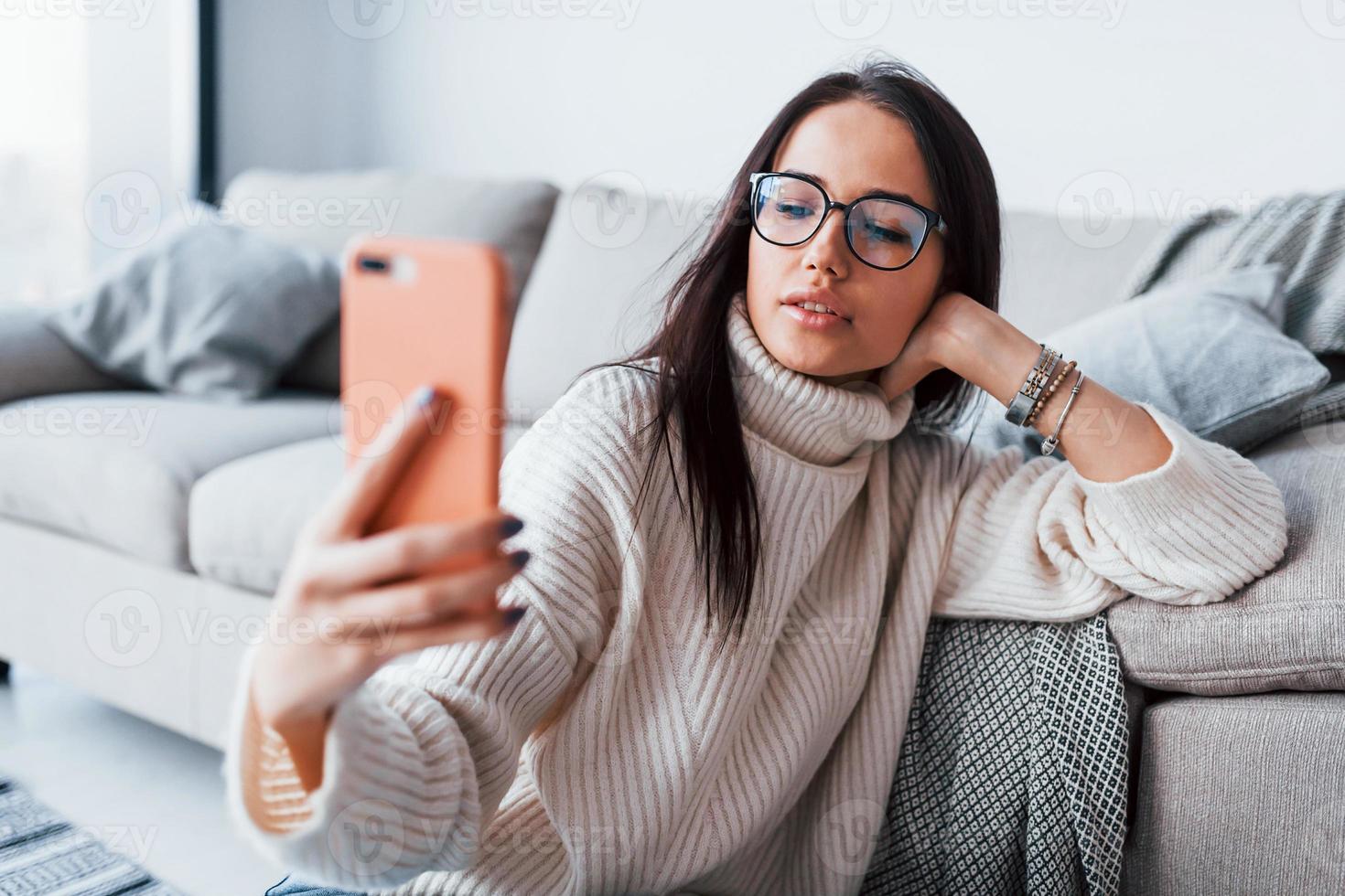 Young beautiful woman in glasses sitting at home alone with phone in hands photo