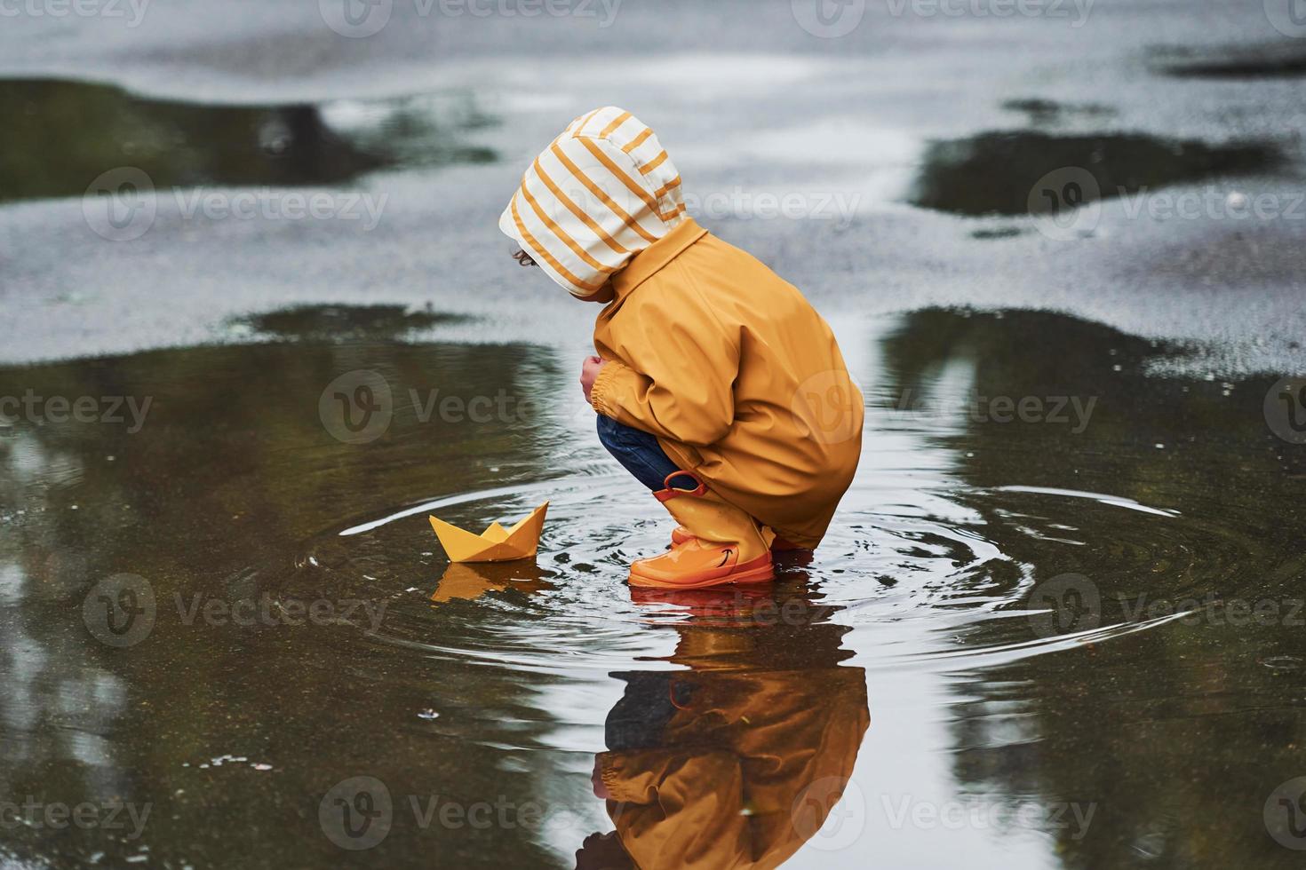 Niño con capa impermeable amarilla y botas jugando al aire libre después de  la lluvia