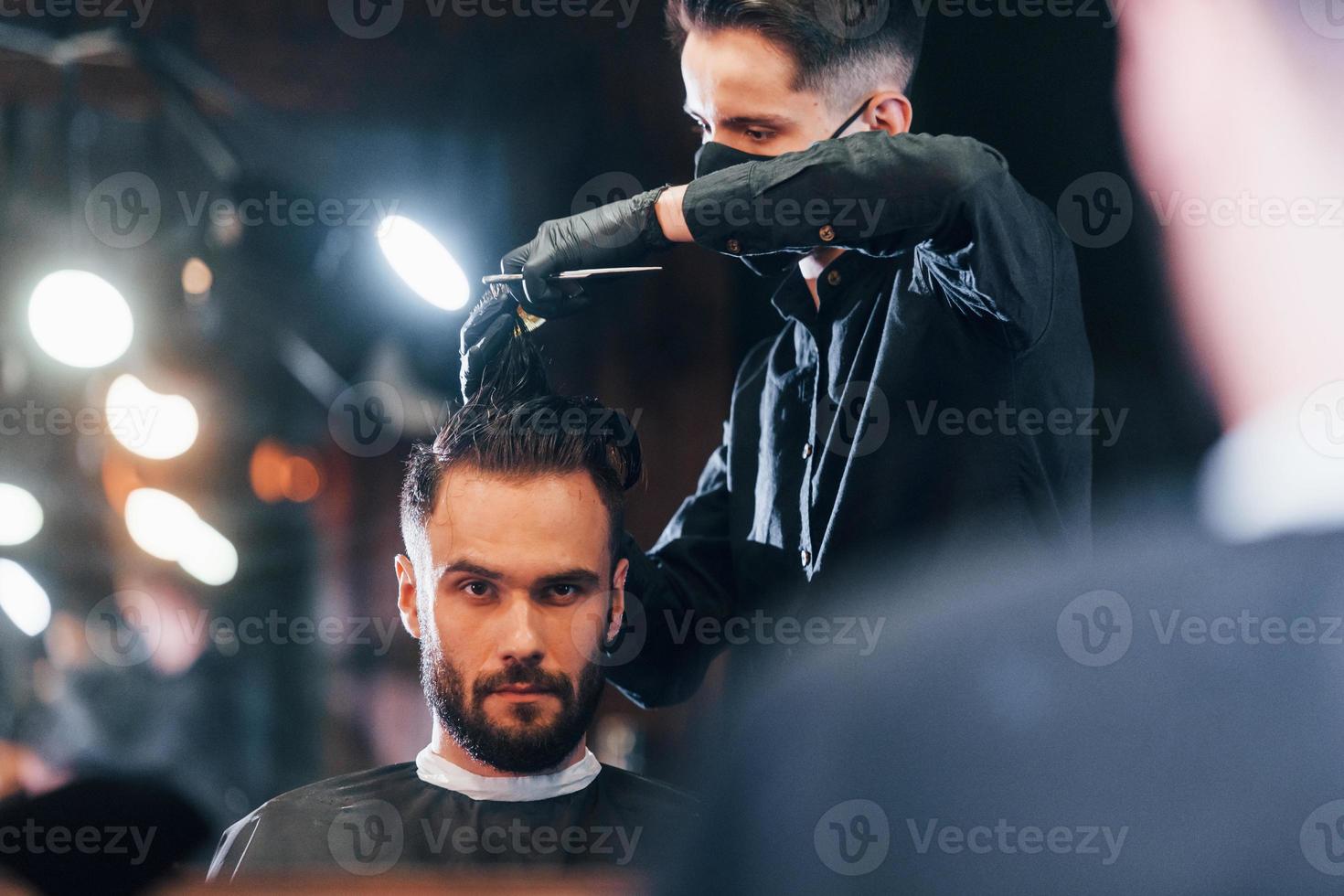 Front view of young bearded man that sitting and getting haircut in barber shop by guy in black protective mask photo
