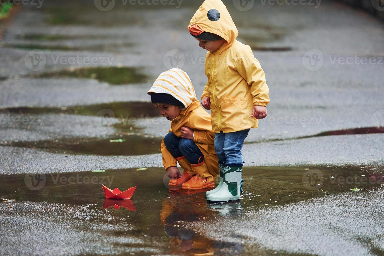 Two kids in yellow waterproof cloaks and boots playing with handmade paper boat outdoors after the rain together photo