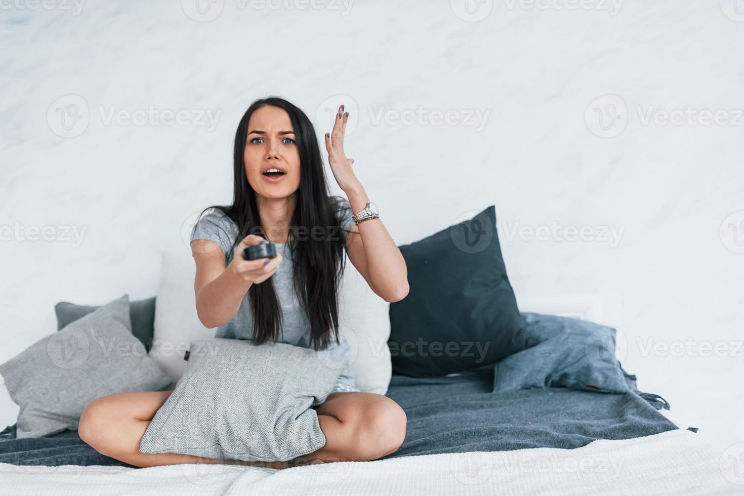 Holding TV remote and watching movie. Young beautiful woman in casual clothes sitting at home alone photo