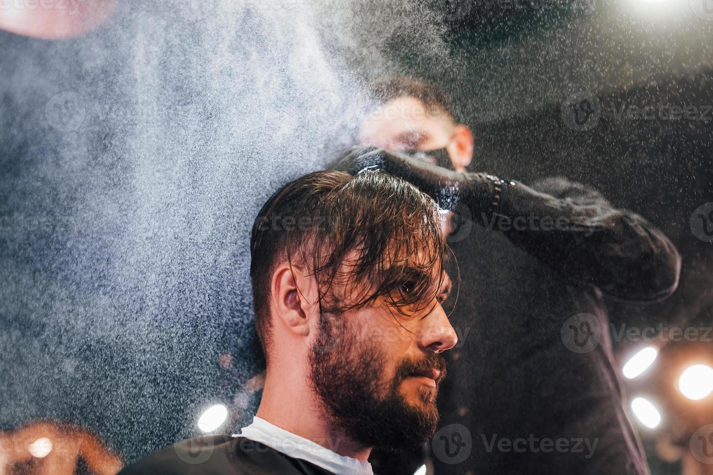 Young bearded man sitting and getting haircut in barber shop by guy in black protective mask photo