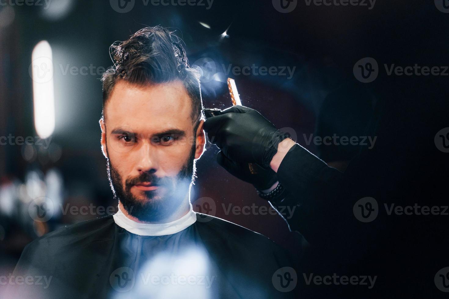 Young bearded man sitting and getting haircut in barber shop photo