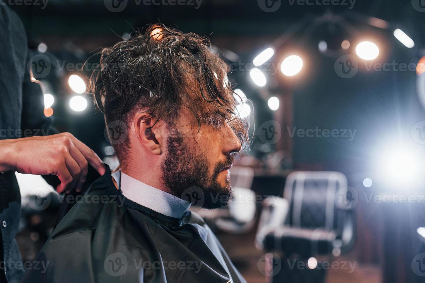 Young bearded man sitting and getting haircut in barber shop photo