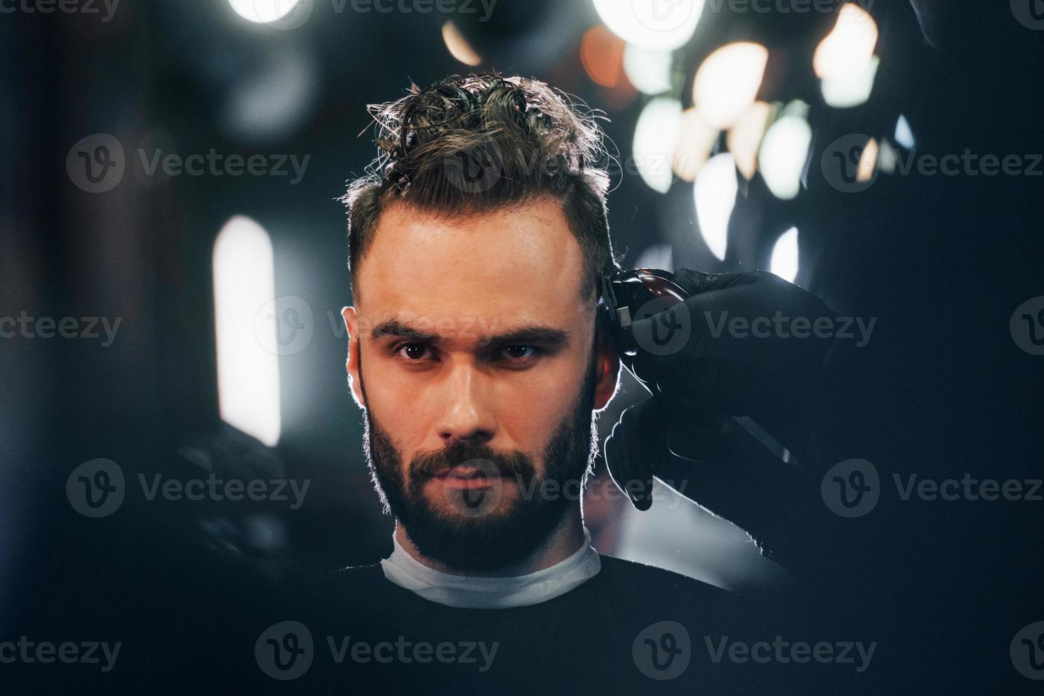 Young bearded man sitting and getting haircut in barber shop photo