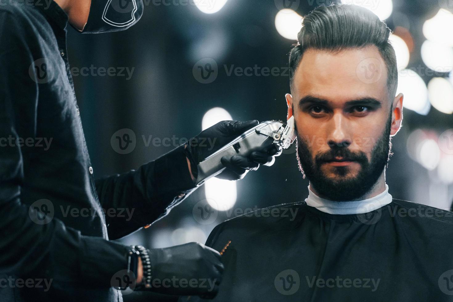 Young man with stylish hairstyle sitting and getting his beard shaved in barber shop photo