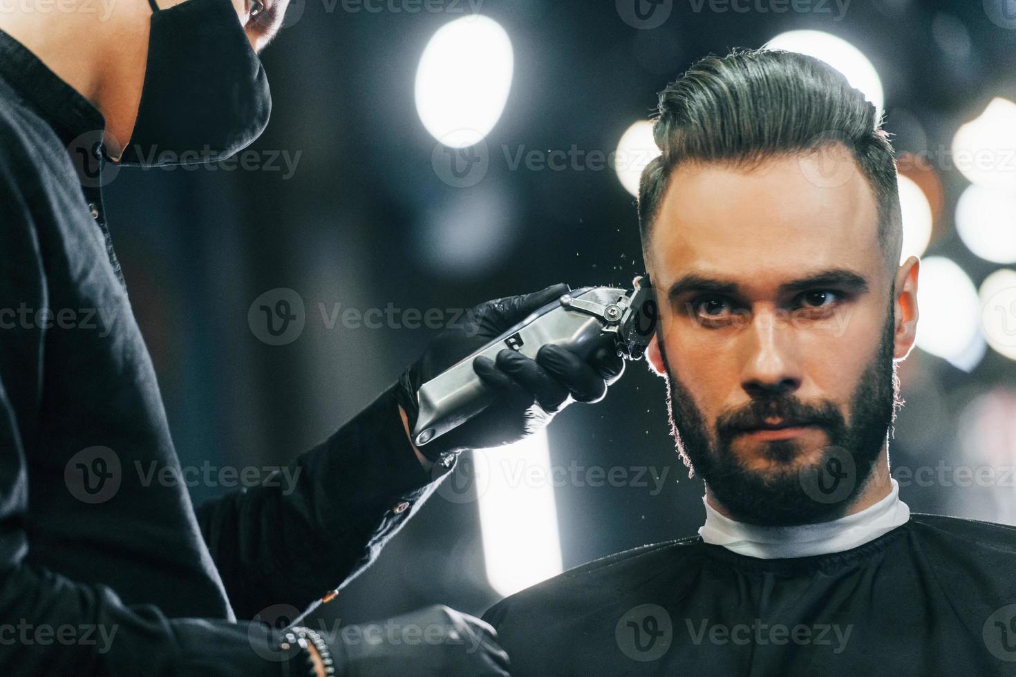 Young man with stylish hairstyle sitting and getting his beard shaved in barber shop photo