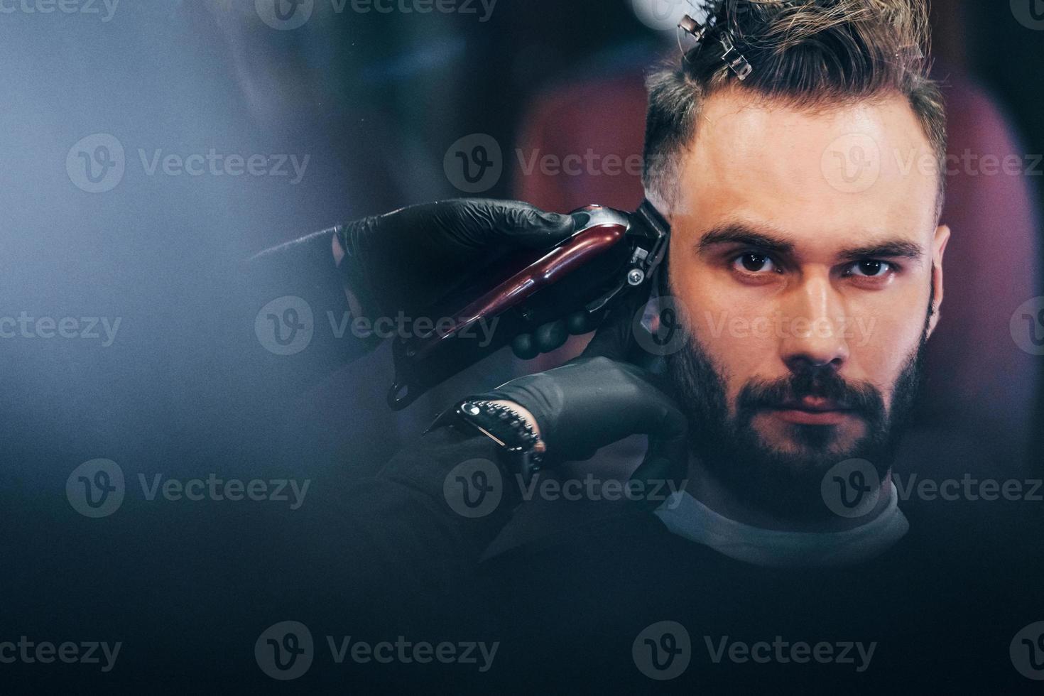 Young bearded man sitting and getting haircut in barber shop photo