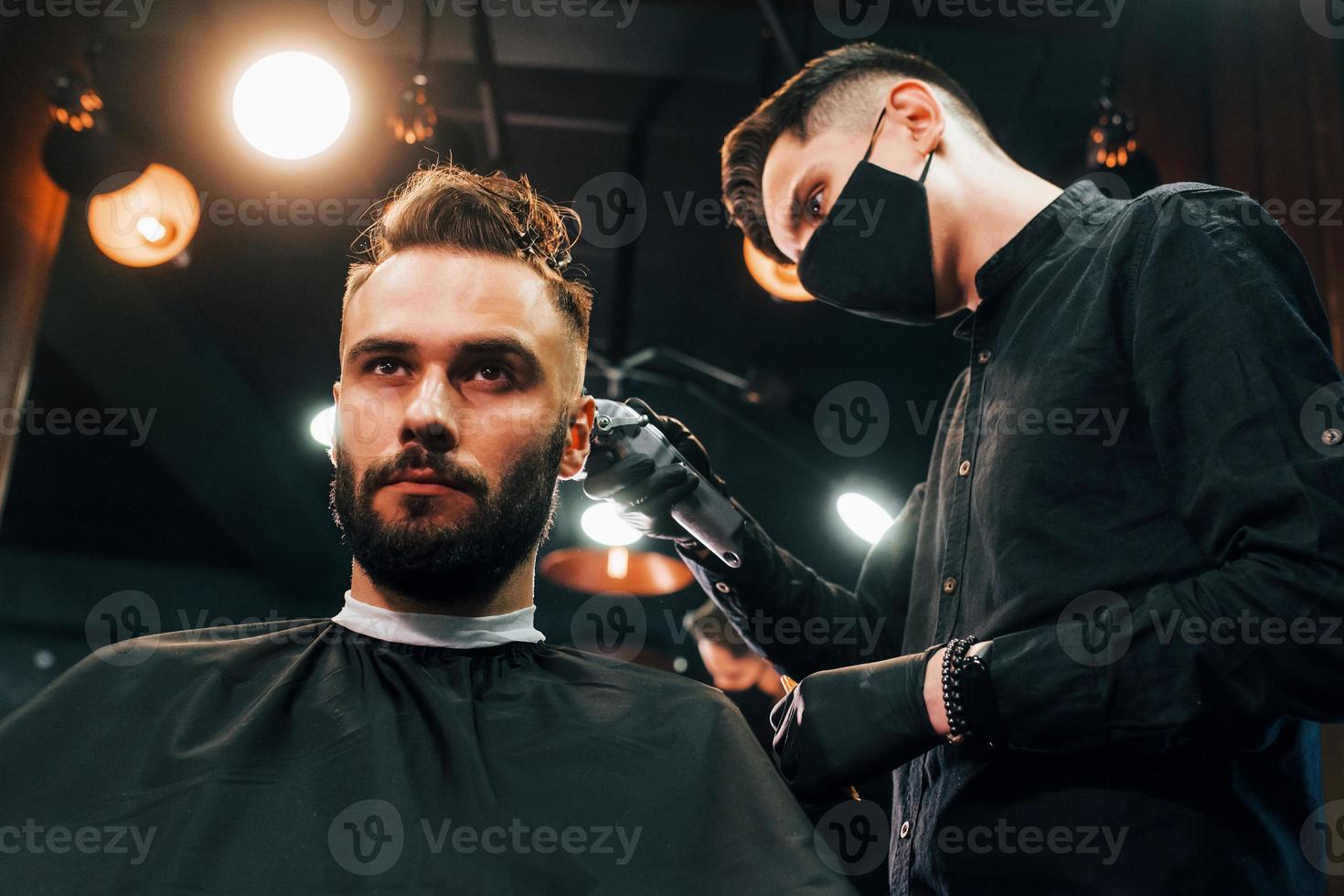 Front view of young bearded man that sitting and getting haircut in barber shop by guy in black protective mask photo