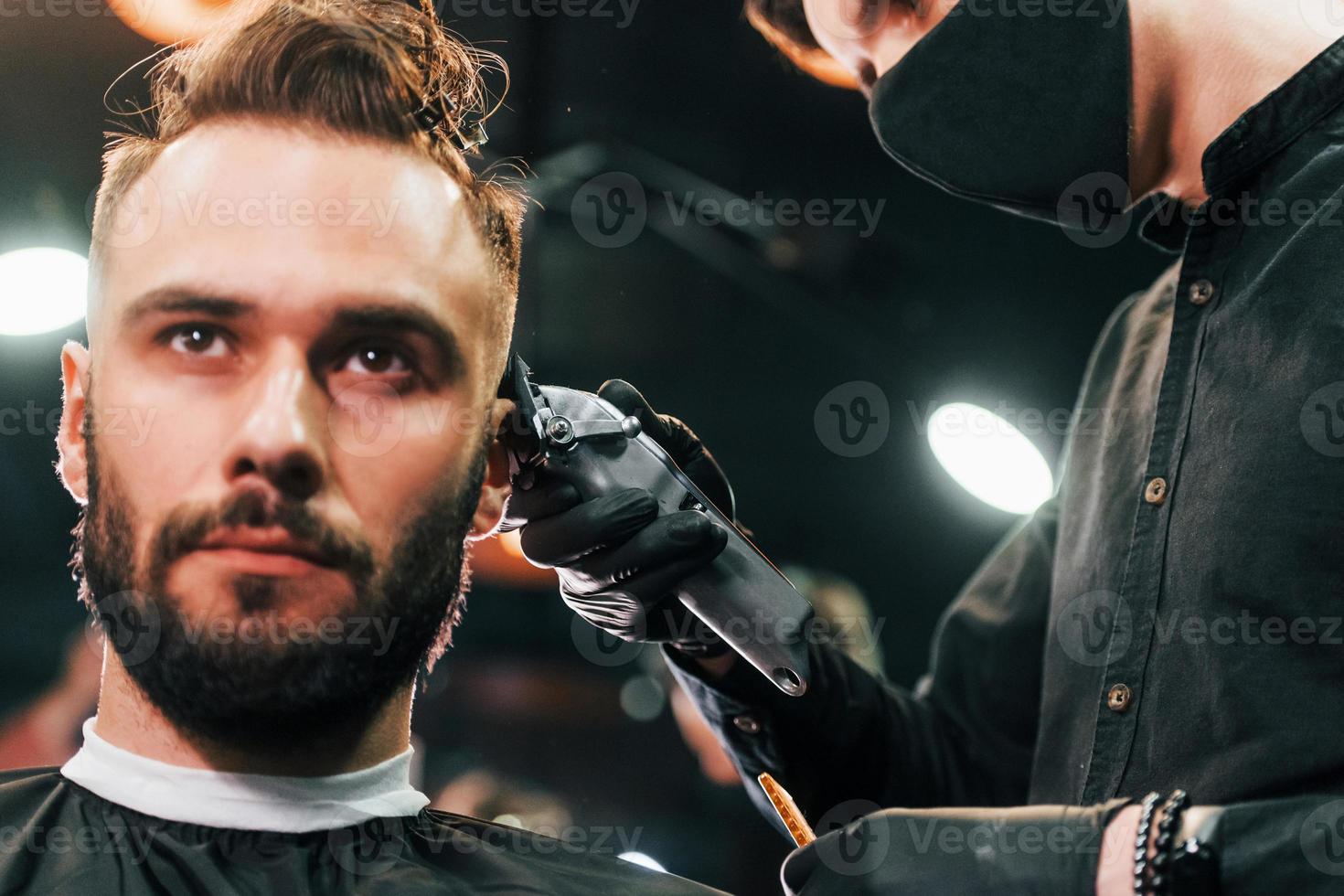 Front view of young bearded man that sitting and getting haircut in barber shop by guy in black protective mask photo