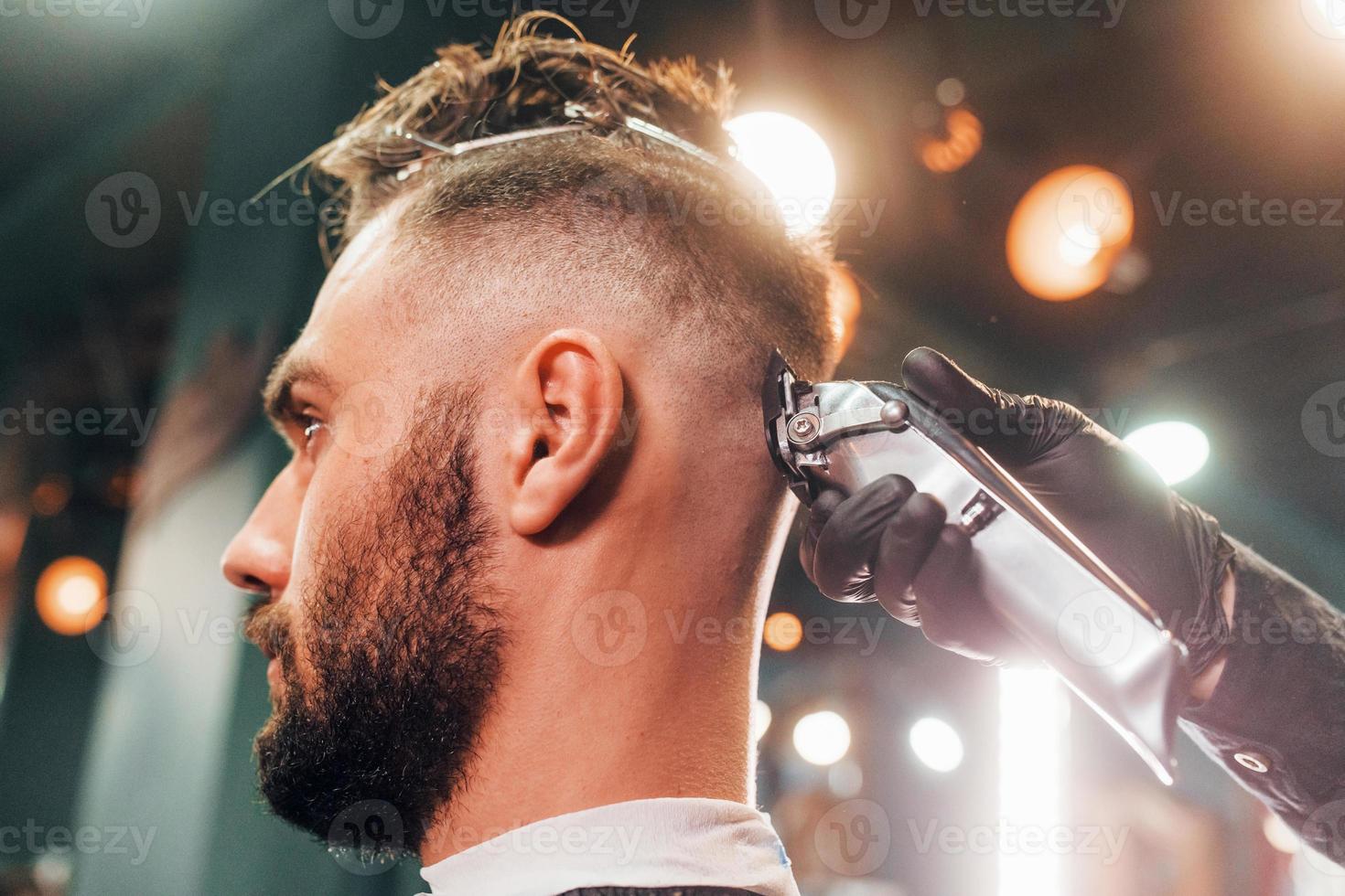 Close up view of young bearded man that sitting and getting haircut in barber shop photo