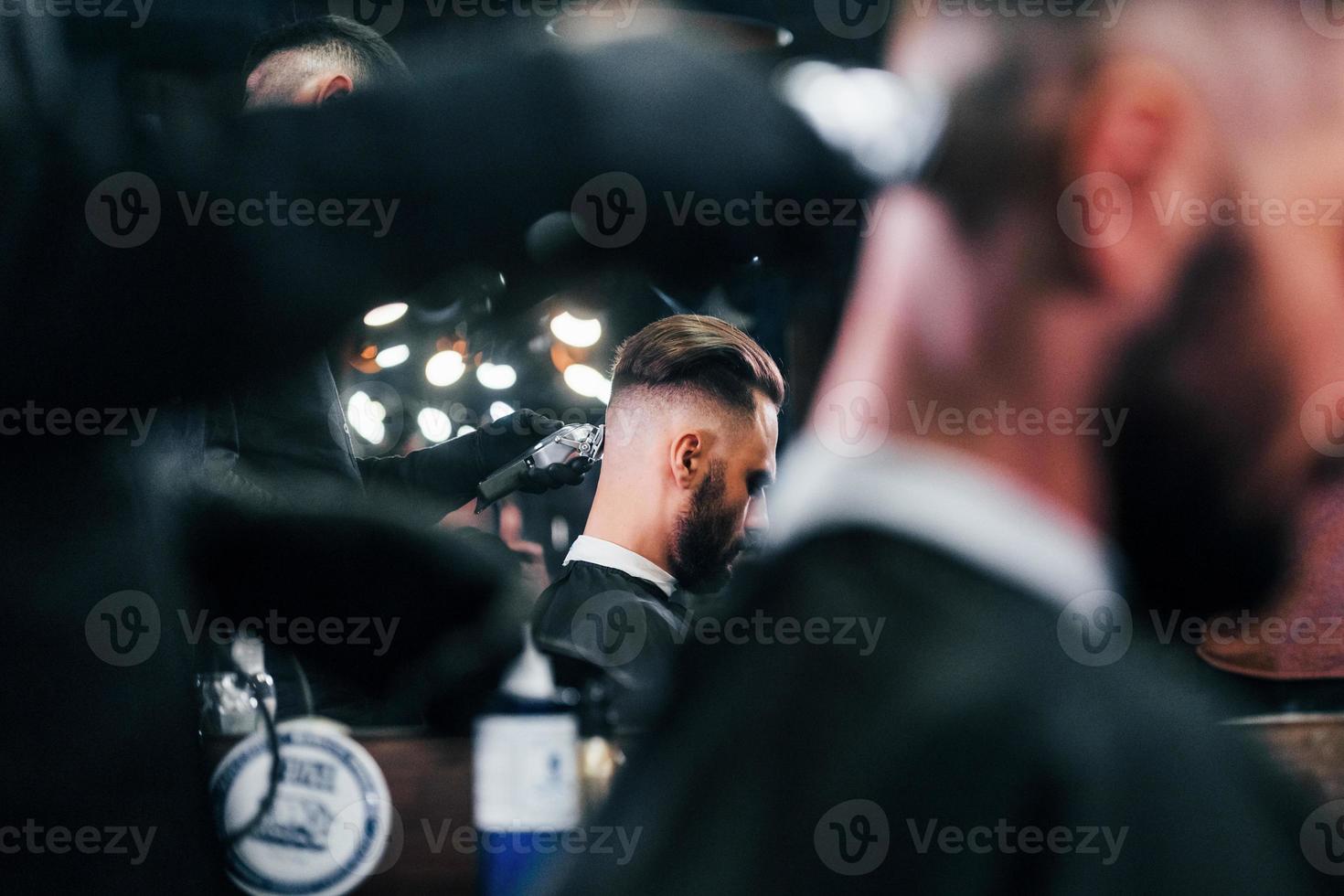 Young bearded man sitting and getting haircut in barber shop photo