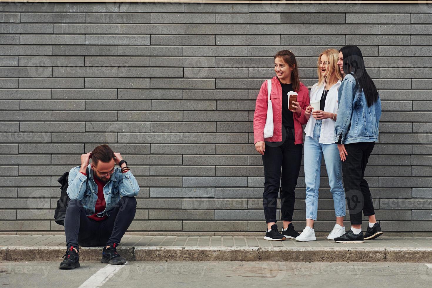 Three women standing and laughing at guy that sitting outdoors near building at daytime photo