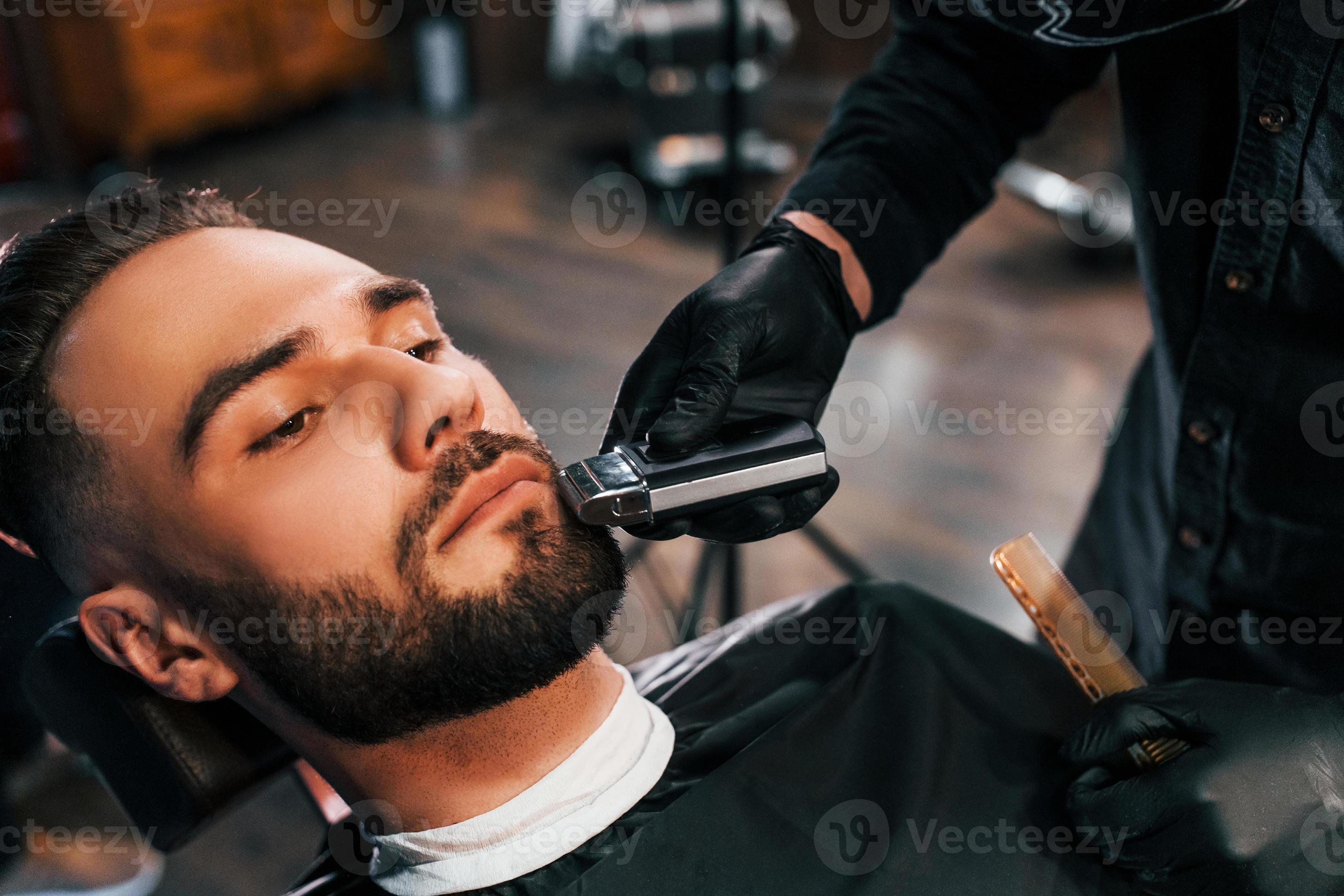 Young man with stylish hairstyle sitting and getting his beard shaved in barber shop 15291399
