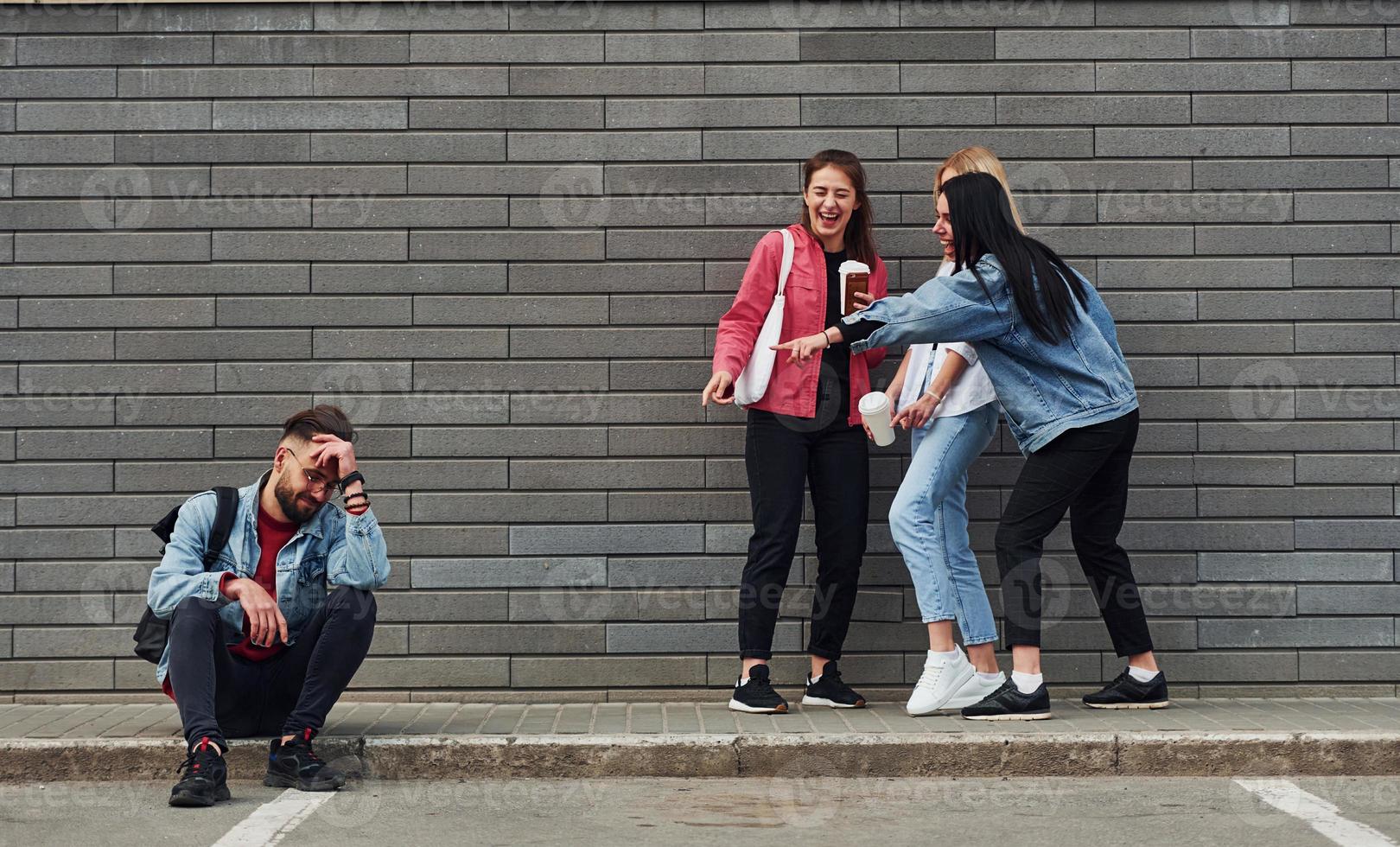 Three women standing and laughing at guy that sitting outdoors near building at daytime photo