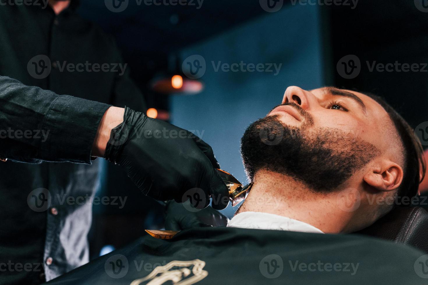 Young man with stylish hairstyle sitting and getting his beard shaved in barber shop photo