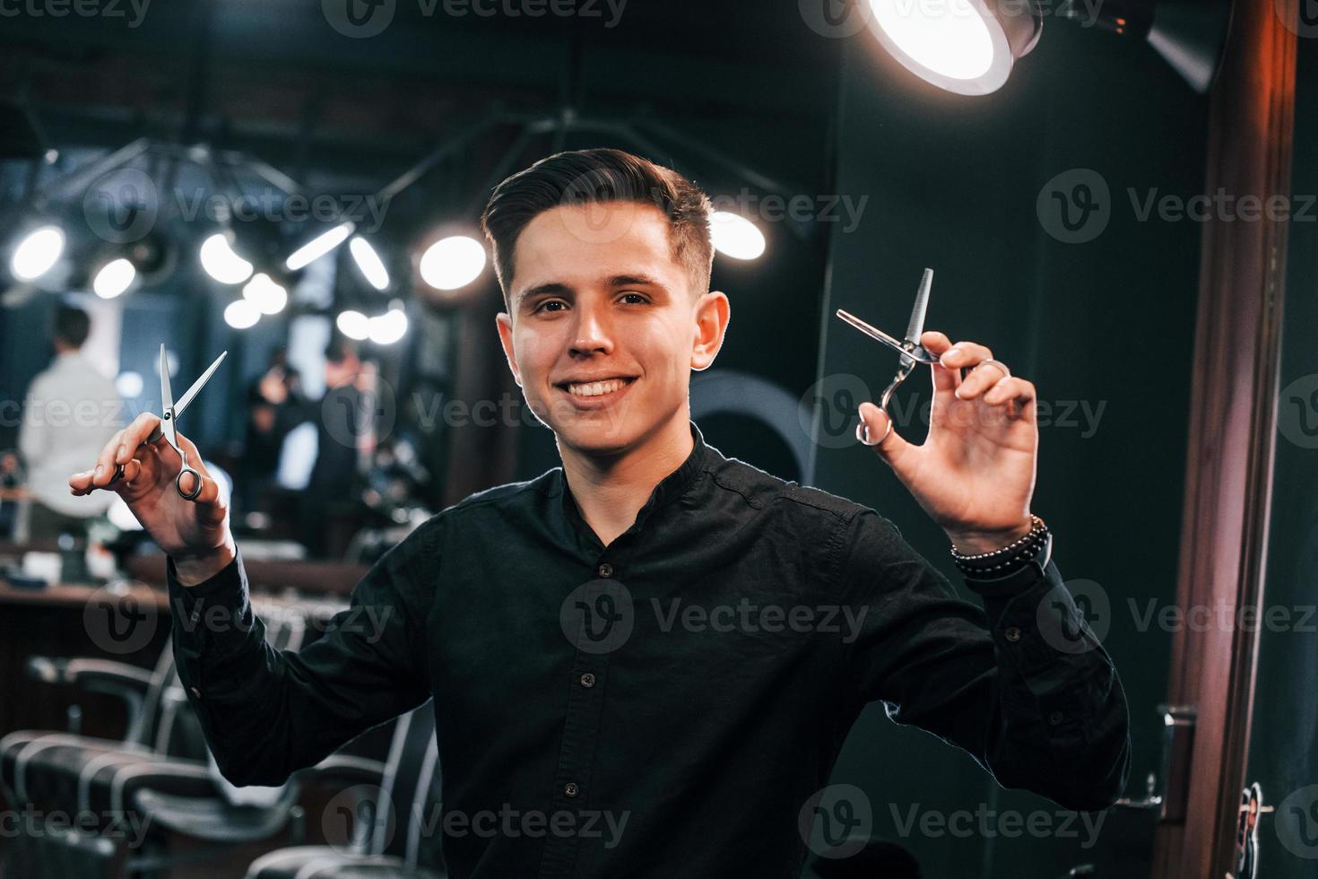 Portrait of young guy in black clothes that standing indoors in barber shop and holding scissors photo