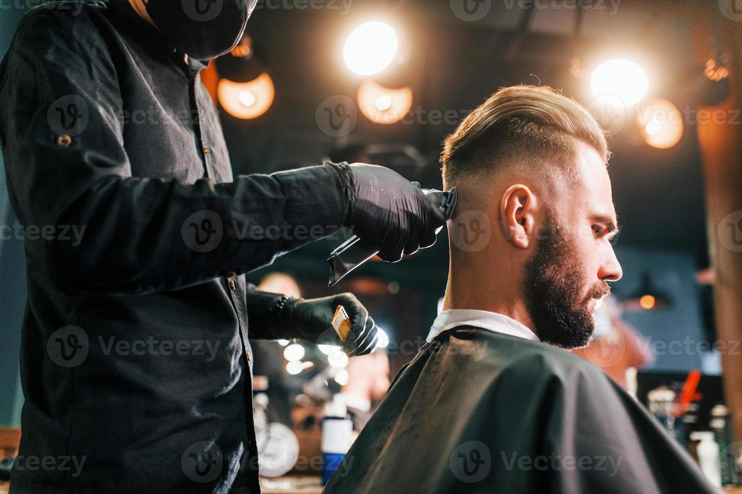 joven barbudo sentado y cortándose el pelo en la barbería por un tipo con máscara protectora negra foto