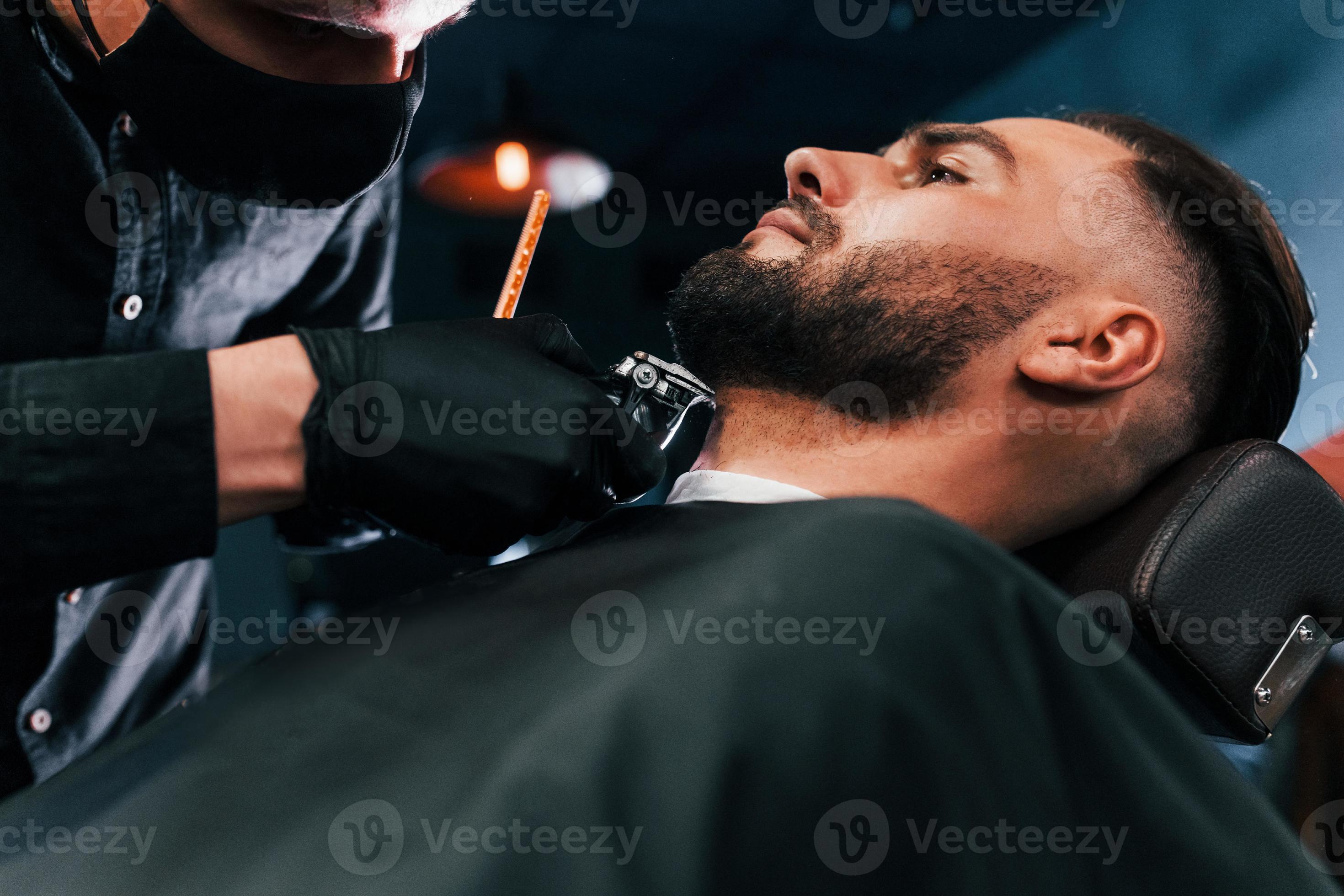joven con peinado elegante sentado y afeitándose la barba por un tipo
