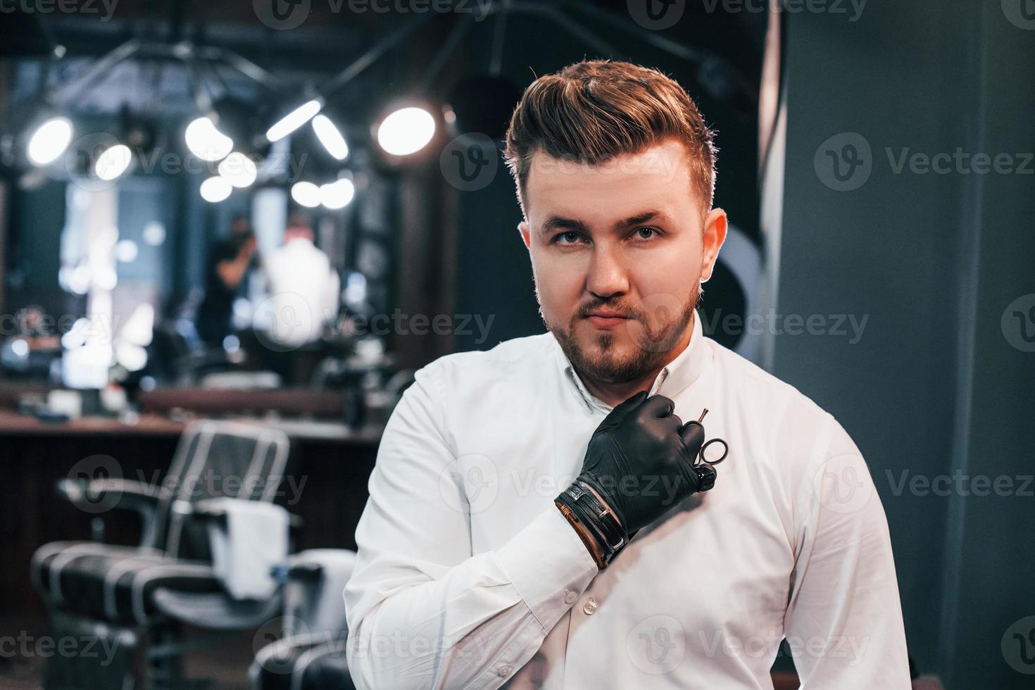 Portrait of young guy in white clothes and black gloves that standing indoors in barber shop and holding scissors photo