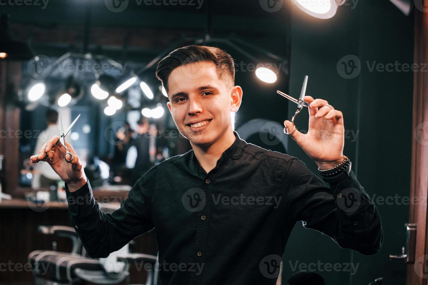 retrato de un joven vestido de negro que está parado adentro en una barbería y sosteniendo unas tijeras foto