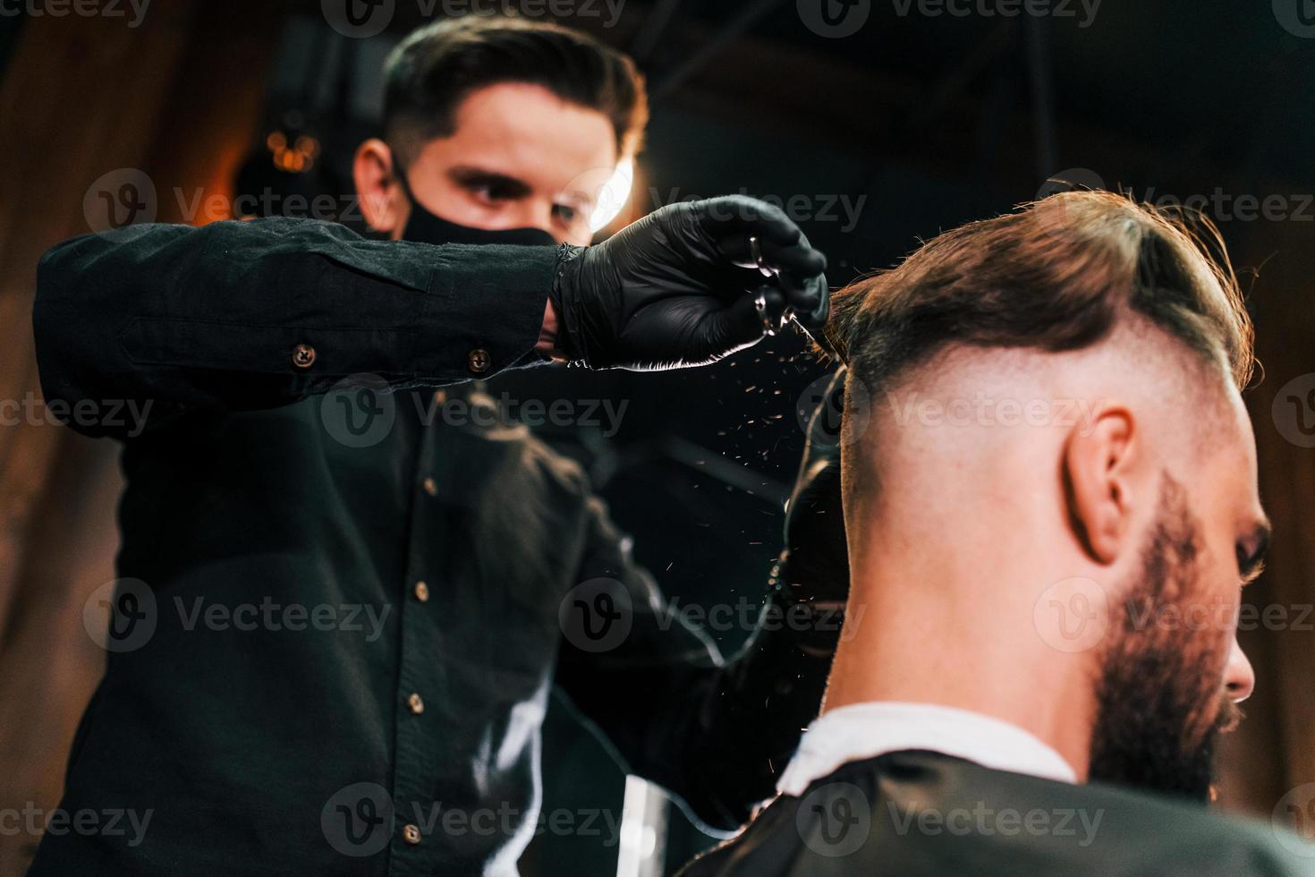 Young bearded man sitting and getting haircut in barber shop by guy in black protective mask photo