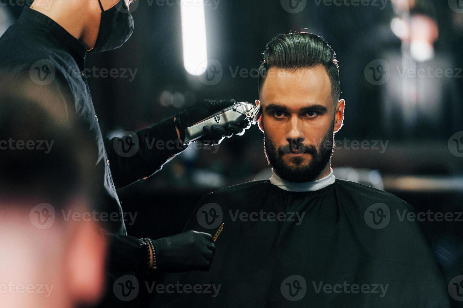 Young man with stylish hairstyle sitting and getting his beard shaved in barber shop photo