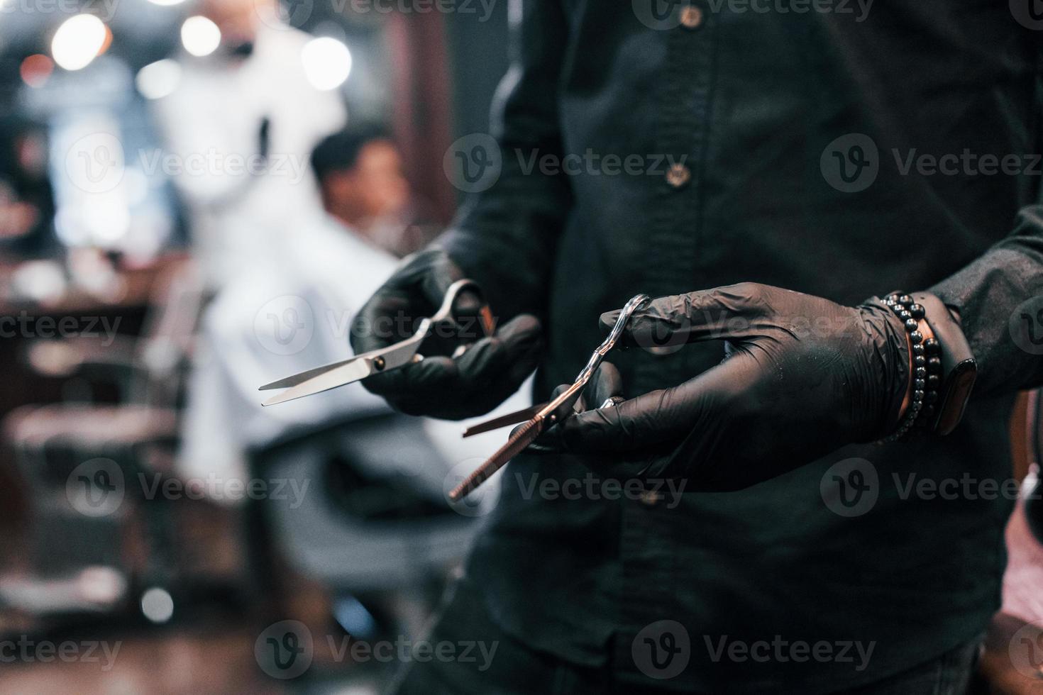 vista de cerca de un tipo vestido de negro que está parado adentro en una barbería y sosteniendo unas tijeras foto