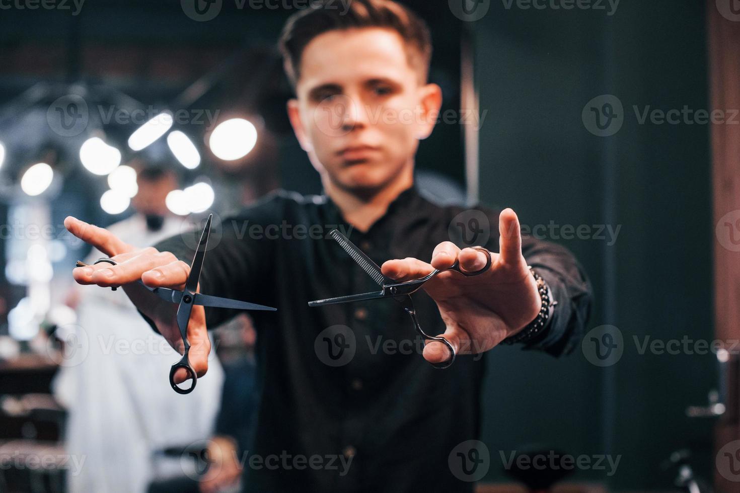 Portrait of young guy in black clothes that standing indoors in barber shop and holding scissors photo