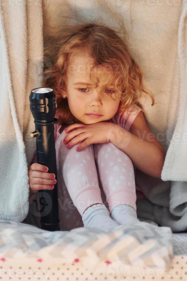Sitting under blanket with flashlight. Cute little girl in casual clothes is indoors at home at daytime photo