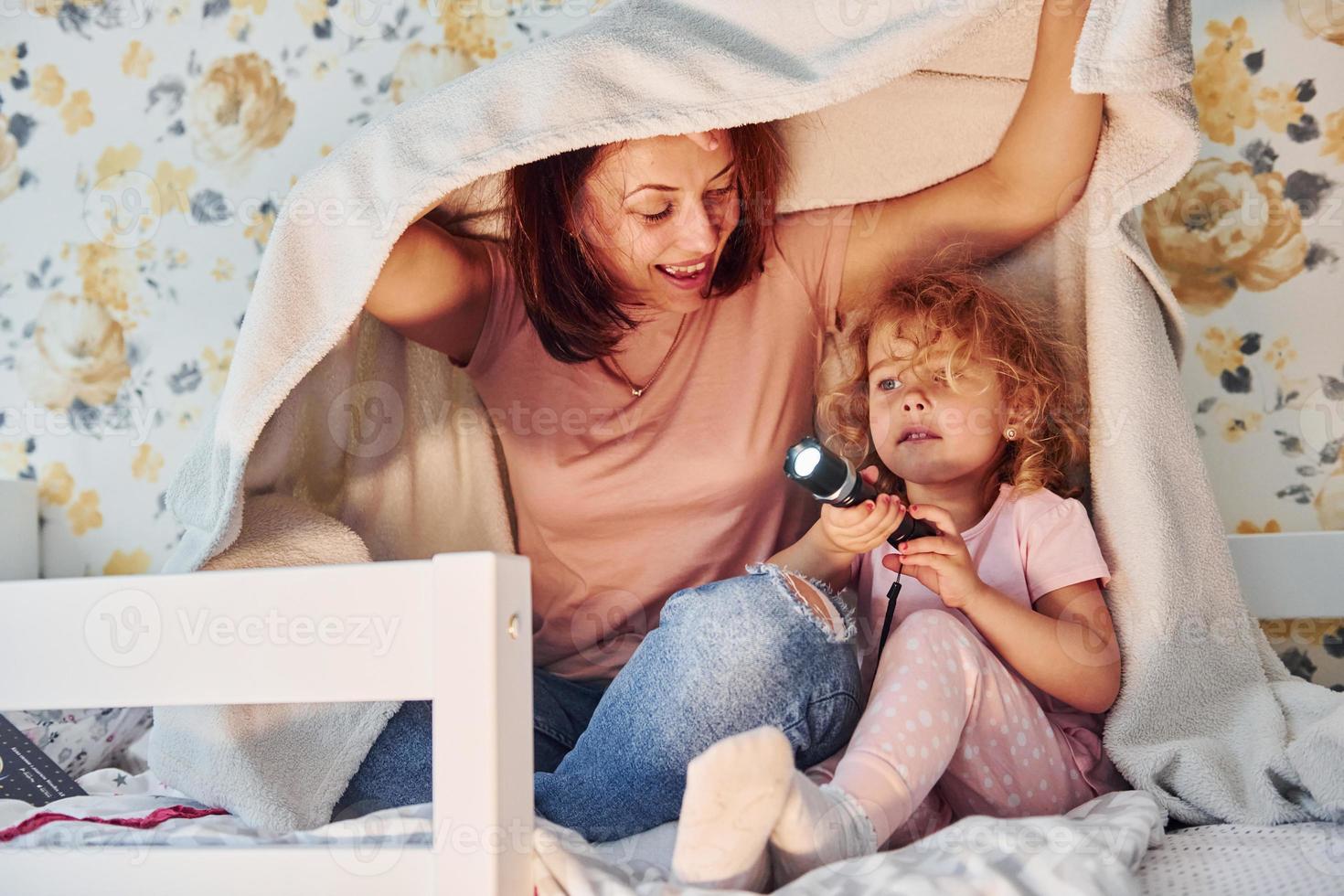 Sitting under blanket with flashlight. Young mother with her little daughter in casual clothes together indoors at home photo