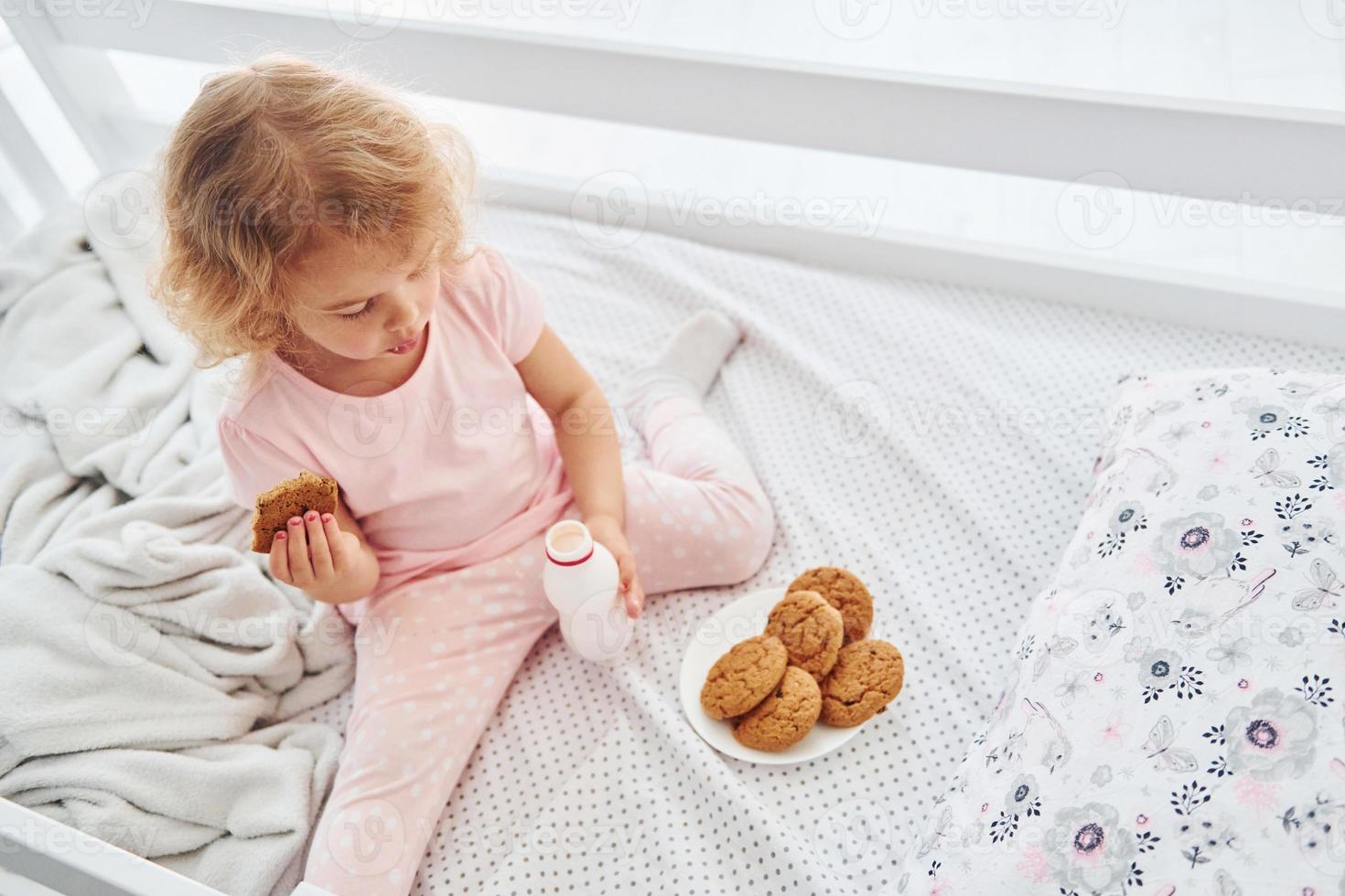 Having a breakfast by eating cookies. Cute little girl in casual clothes is indoors at home at daytime photo