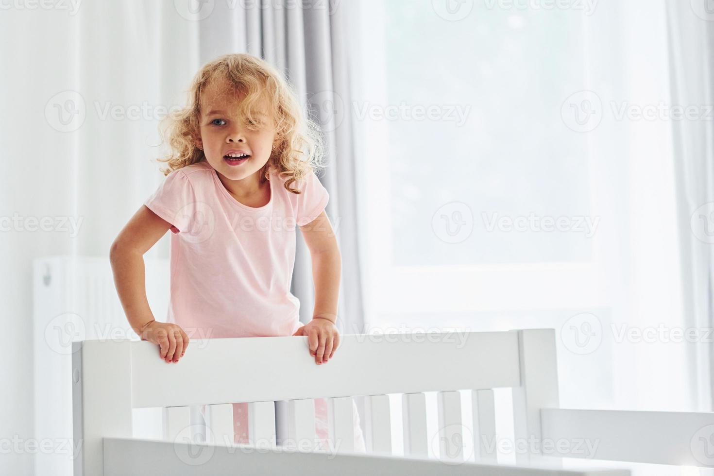 Standing on the bed. Cute little girl in casual clothes is indoors at home at daytime photo