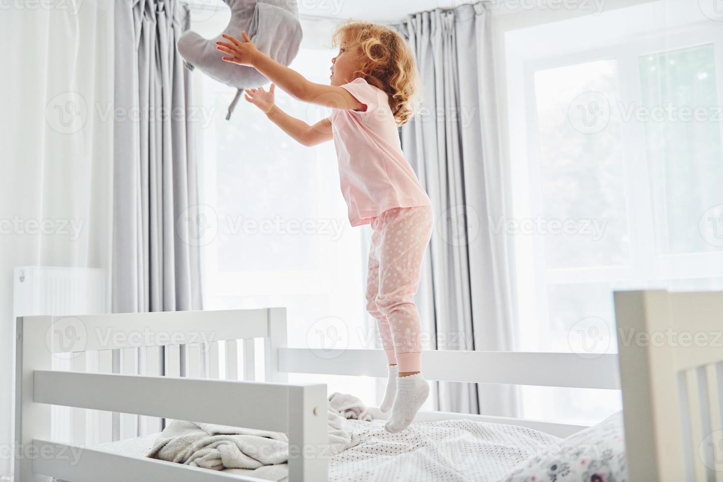 Playing with pillow by throwing it. Cute little girl in casual clothes is indoors at home at daytime photo