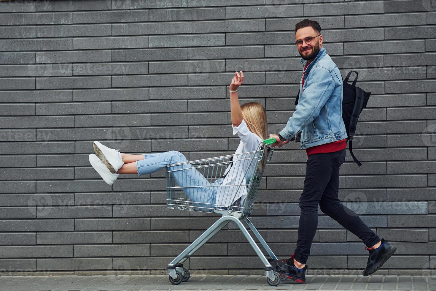 divirtiéndose y montando carrito de compras. joven elegante con mujer en ropa casual al aire libre juntos. concepción de la amistad o las relaciones foto