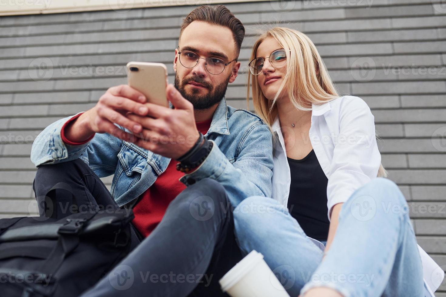 Using phone. Young stylish man with woman in casual clothes sitting outdoors together. Conception of friendship or relationships photo