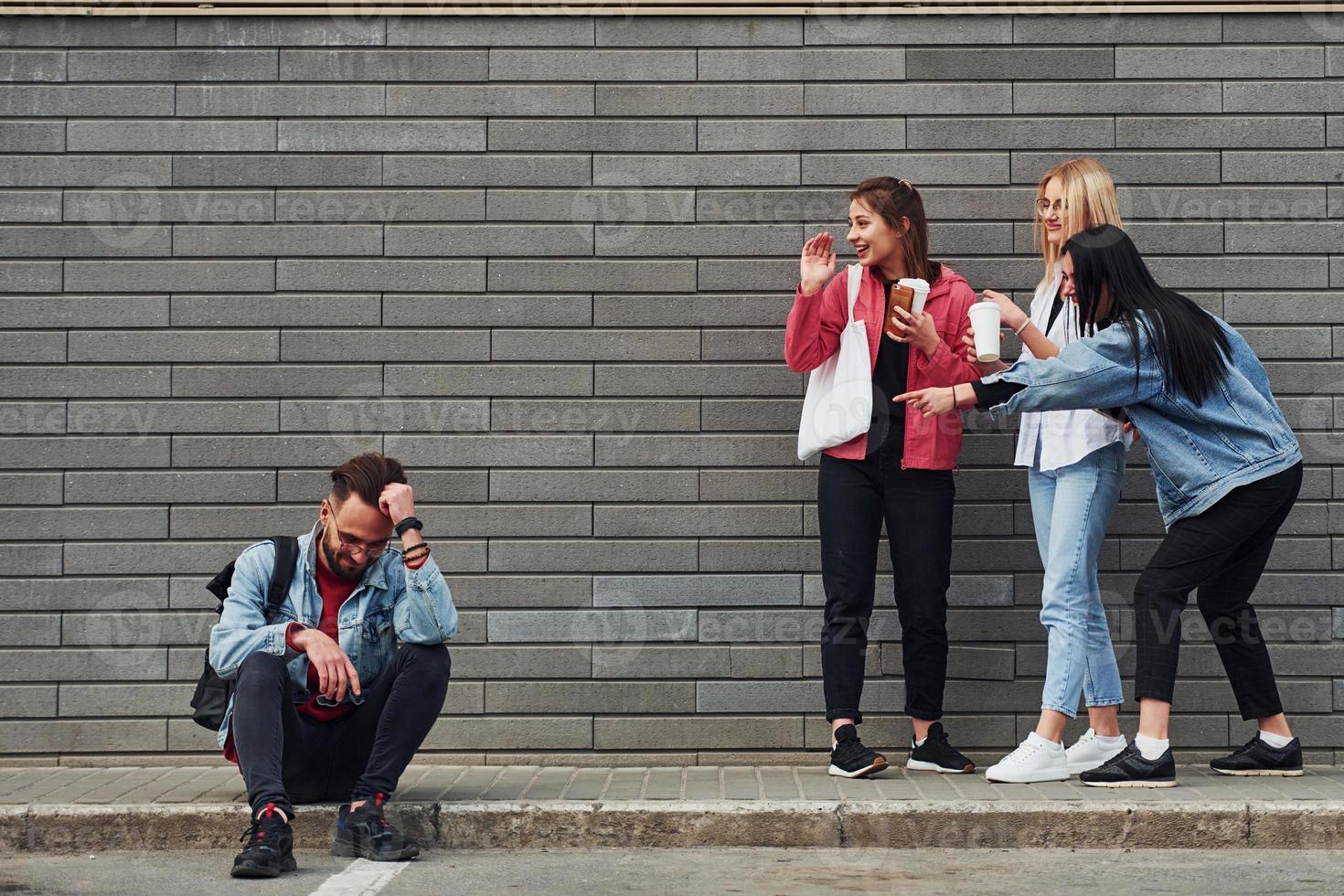 Three women standing and laughing at guy that sitting outdoors near building at daytime photo