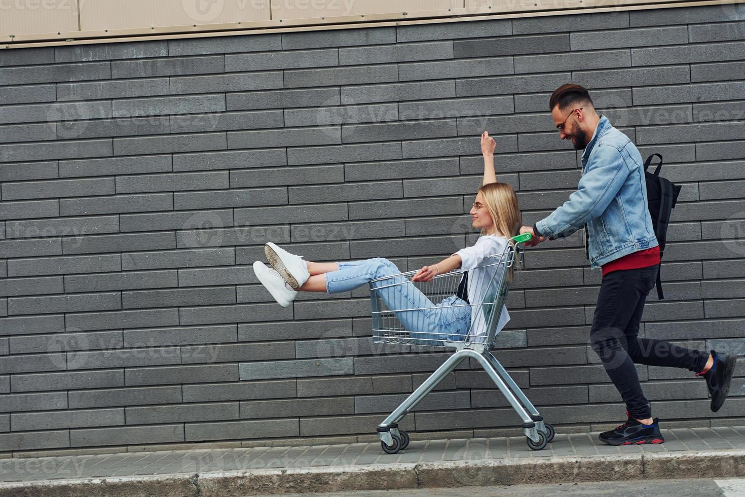 Having fun and riding shopping cart. Young stylish man with woman in casual clothes outdoors together. Conception of friendship or relationships photo