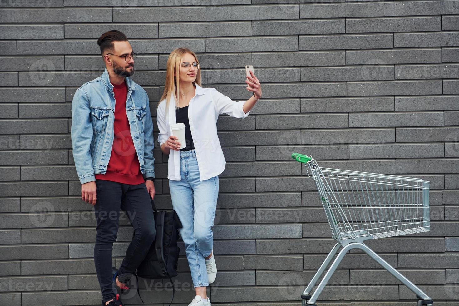 With shopping cart. Young stylish man with woman in casual clothes outdoors together. Conception of friendship or relationships photo
