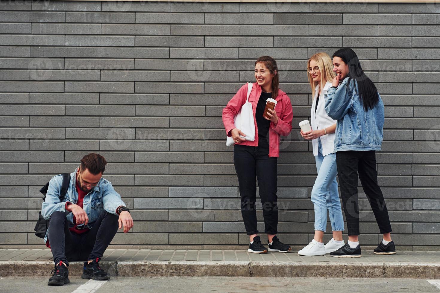 Three women standing and laughing at guy that sitting outdoors near building at daytime photo