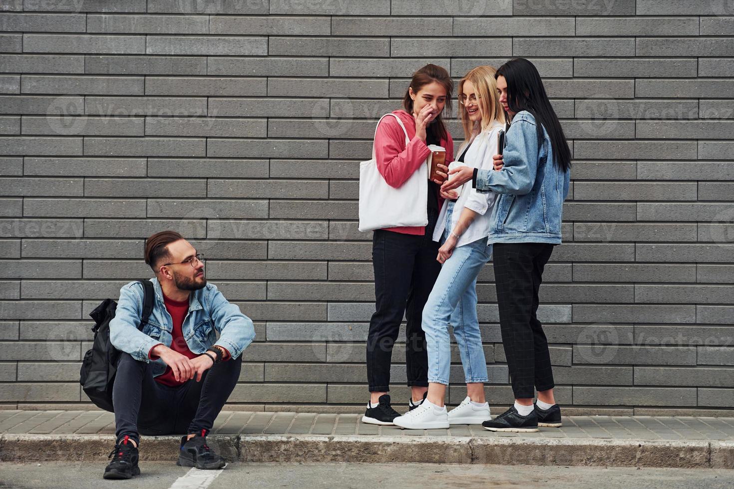 Three women standing and laughing at guy that sitting outdoors near building at daytime photo