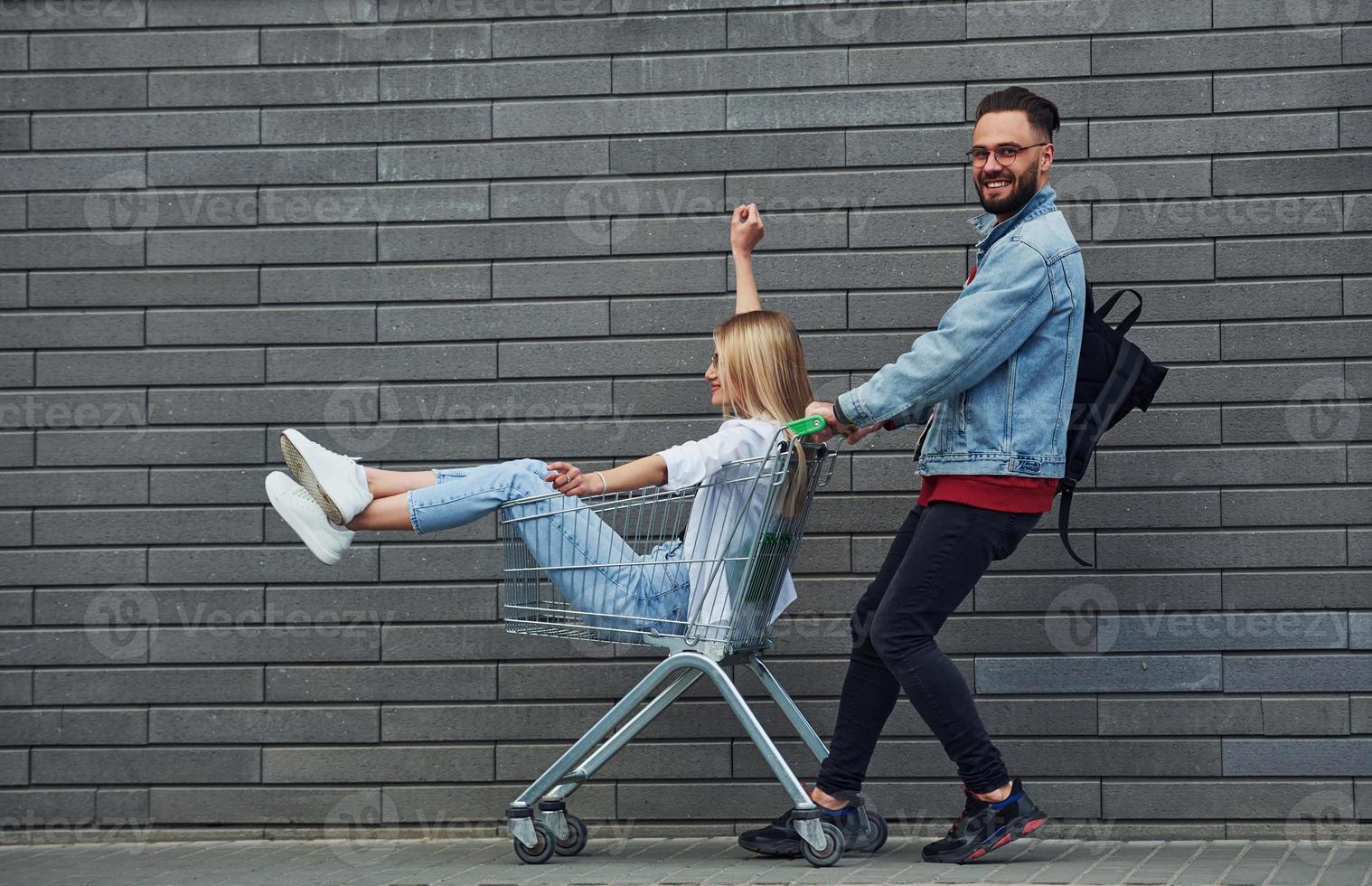 Having fun and riding shopping cart. Young stylish man with woman in casual clothes outdoors together. Conception of friendship or relationships photo