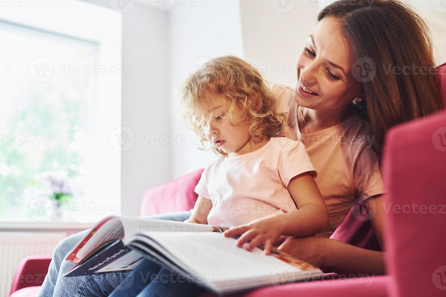 leyendo un libro grande. joven madre con su pequeña hija vestida de forma informal juntas en casa foto