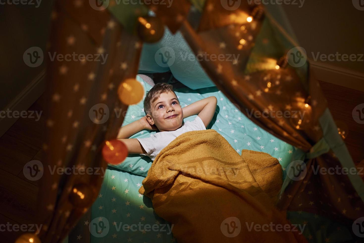 Young boy in casual clothes lying down in tent at evening time photo