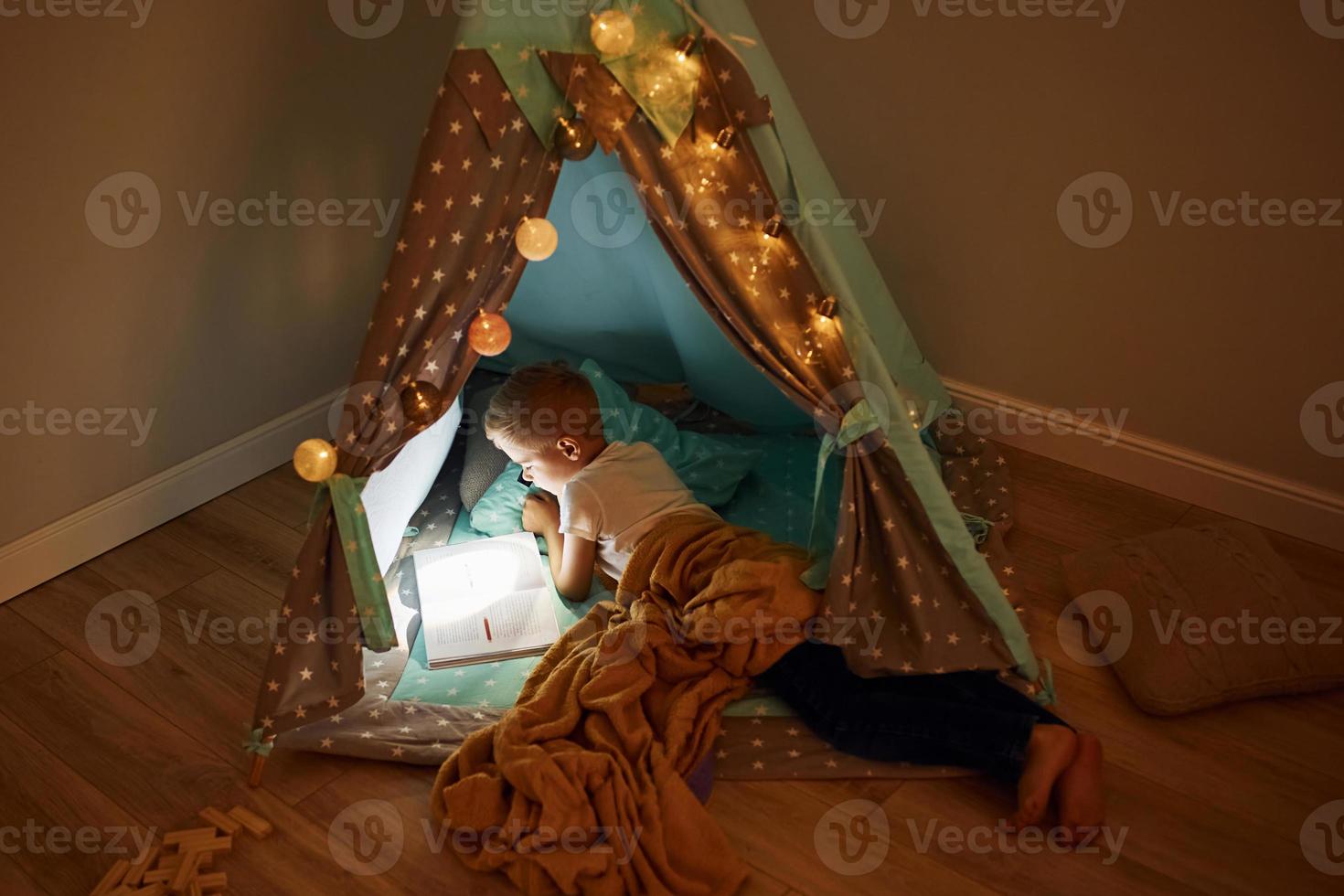 Reading book and using flashlight. Young boy in casual clothes lying down near tent at evening time photo