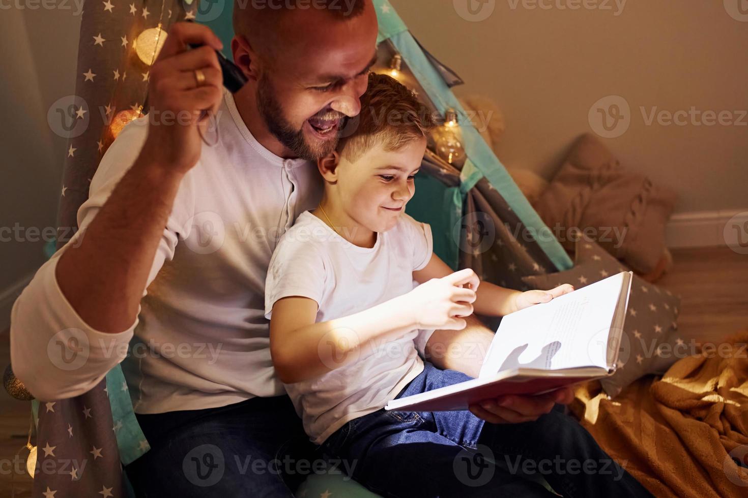 Reading book and using flashlight. Father and his young young spending time together indoors near tent photo