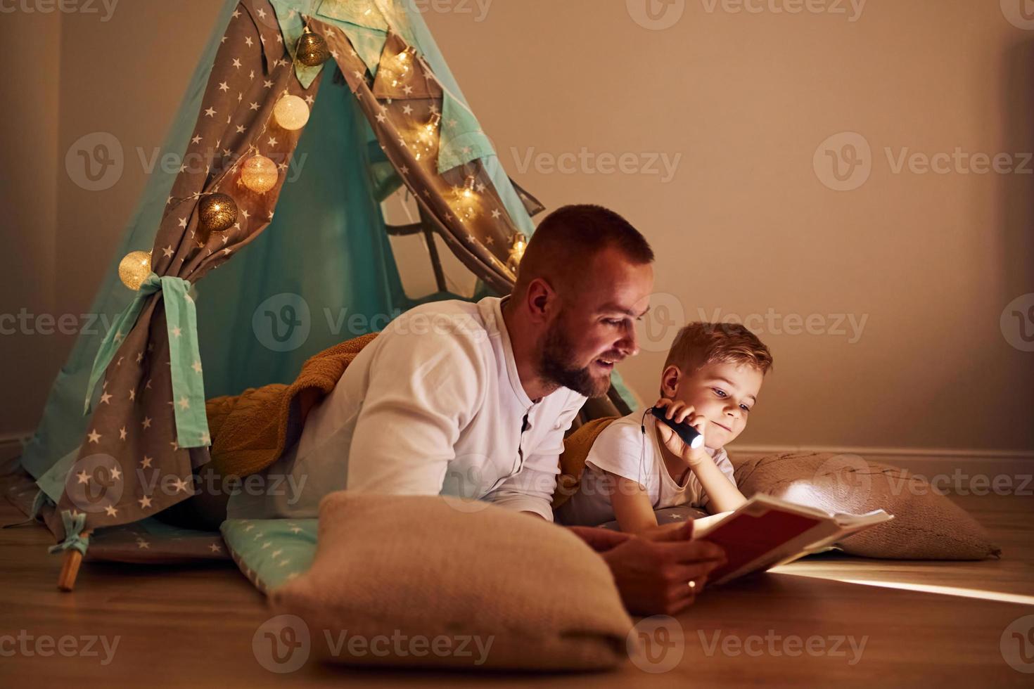 Reading book and using flashlight. Father and his young young spending time together indoors near tent photo
