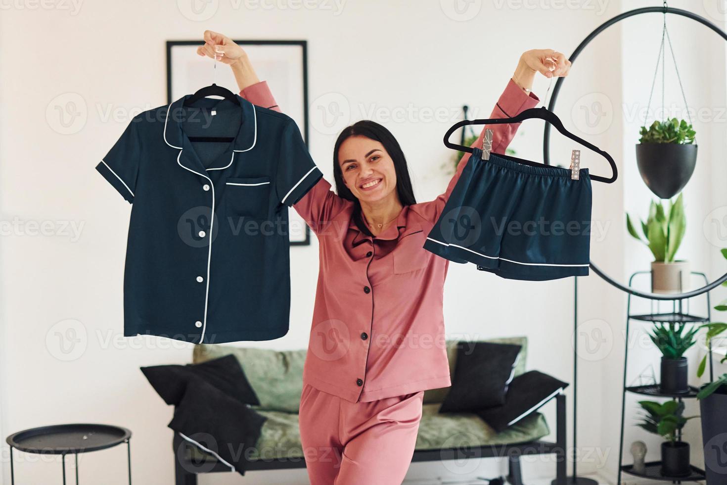 sosteniendo ropa azul en las manos. mujer joven alegre en pijama de pie en el interior durante el día foto