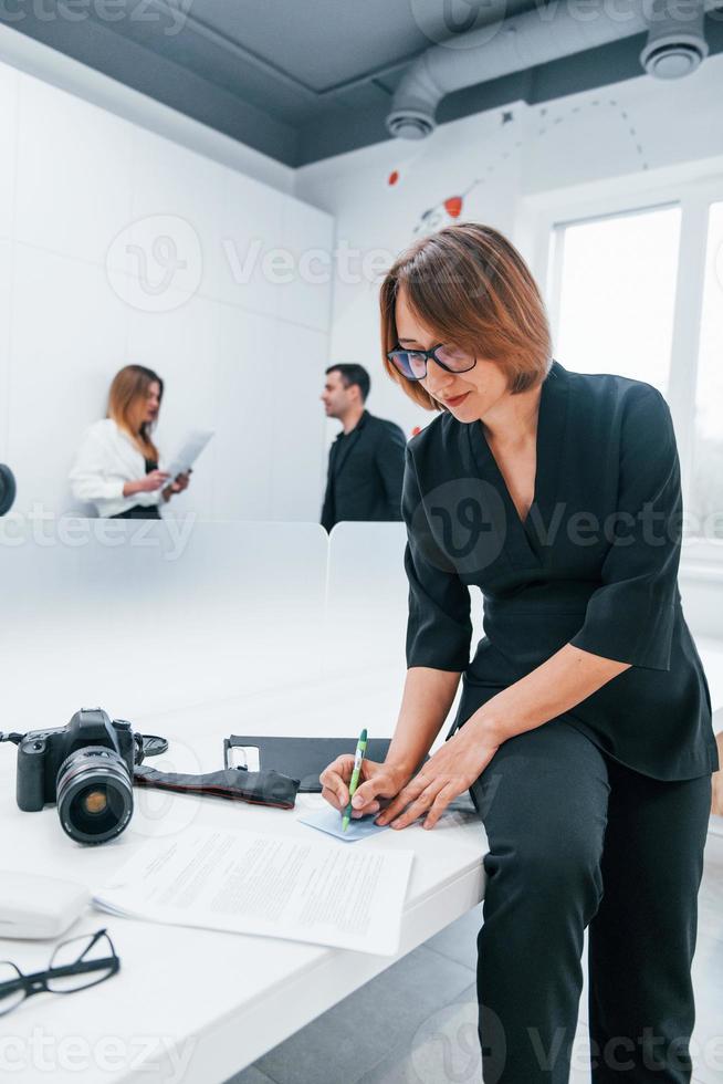 Woman in black formal clothes and with camera in the office. Colleagues behind photo